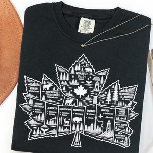 May include: A black crew neck t-shirt featuring a large white graphic of a maple leaf divided into sections representing Canadian provinces and territories, each containing illustrations of local landmarks, wildlife, and city names.