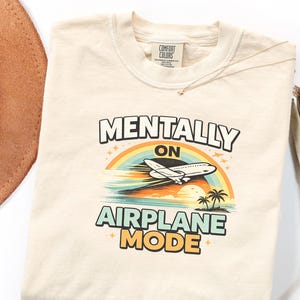 Puede incluir: Camiseta color crema con las palabras "MENTALLY ON AIRPLANE MODE" en una fuente divertida. El dise&ntilde;o presenta un arco&iacute;ris, un avi&oacute;n, palmeras y el sol. La camiseta es de material suave y tiene cuello redondo.
