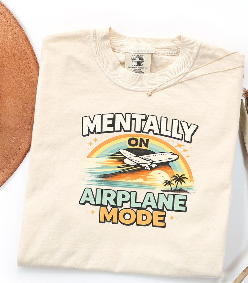 Puede incluir: Camiseta color crema con las palabras "MENTALLY ON AIRPLANE MODE" en una fuente divertida. El dise&ntilde;o presenta un arco&iacute;ris, un avi&oacute;n, palmeras y el sol. La camiseta es de material suave y tiene cuello redondo.