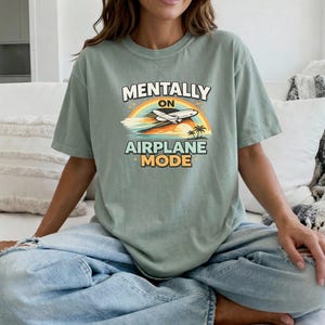 Puede incluir: Camiseta verde salvia con el texto "MENTALLY ON AIRPLANE MODE" en un dise&ntilde;o retro. El gr&aacute;fico incluye un arco&iacute;ris, un avi&oacute;n y palmeras. La camiseta es de corte holgado.