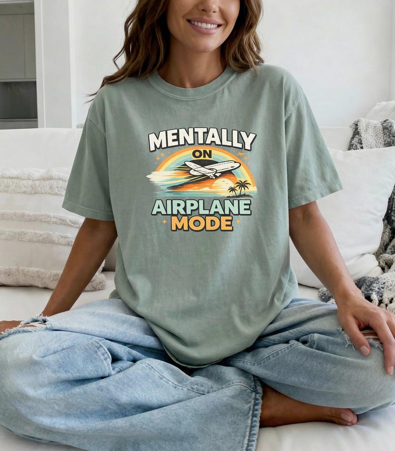 Puede incluir: Camiseta verde salvia con el texto "MENTALLY ON AIRPLANE MODE" en un dise&ntilde;o retro. El gr&aacute;fico incluye un arco&iacute;ris, un avi&oacute;n y palmeras. La camiseta es de corte holgado.
