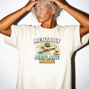 Puede incluir: Camiseta color crema con el texto "MENTALLY ON AIRPLANE MODE" sobre un gr&aacute;fico de un avi&oacute;n volando sobre un arco&iacute;ris, oc&eacute;ano y palmeras. La camiseta es de estilo casual y unisex.