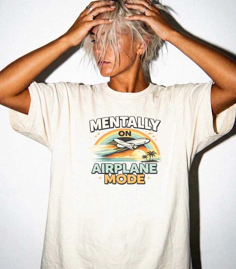 Puede incluir: Camiseta color crema con el texto "MENTALLY ON AIRPLANE MODE" sobre un gr&aacute;fico de un avi&oacute;n volando sobre un arco&iacute;ris, oc&eacute;ano y palmeras. La camiseta es de estilo casual y unisex.