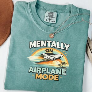 Puede incluir: Camiseta verde azulado con las palabras "MENTALLY ON AIRPLANE MODE" en blanco y amarillo. El dise&ntilde;o incluye un arco&iacute;ris, un avi&oacute;n y palmeras. Un collar plateado con un colgante descansa sobre la camiseta.