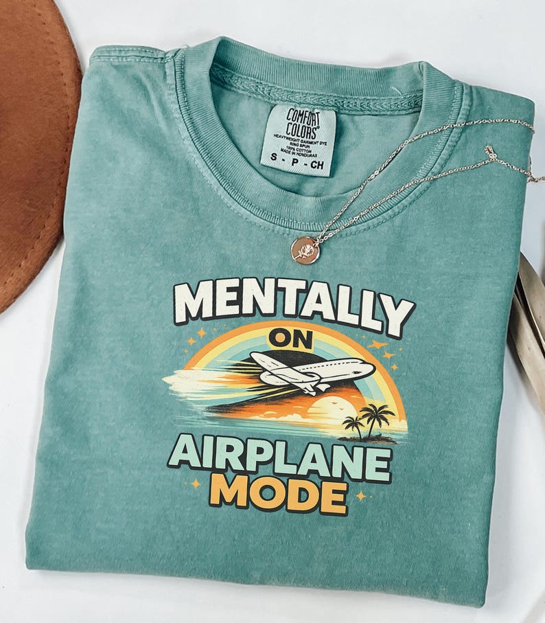 Puede incluir: Camiseta verde azulado con las palabras "MENTALLY ON AIRPLANE MODE" en blanco y amarillo. El dise&ntilde;o incluye un arco&iacute;ris, un avi&oacute;n y palmeras. Un collar plateado con un colgante descansa sobre la camiseta.