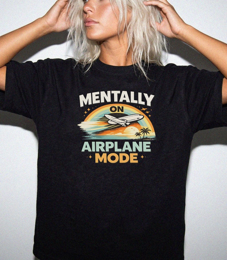 Puede incluir: Camiseta negra con el texto "MENTALLY ON AIRPLANE MODE" sobre un gr&aacute;fico de un avi&oacute;n, arco&iacute;ris, oc&eacute;ano y palmeras. El dise&ntilde;o utiliza colores azul, naranja y verde.
