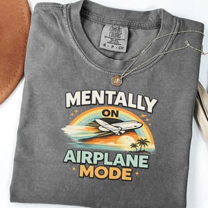 Puede incluir: Camiseta gris con la frase "MENTALLY ON AIRPLANE MODE" en blanco y amarillo. El dise&ntilde;o incluye un arco&iacute;ris, un avi&oacute;n, palmeras y una puesta de sol. Un collar plateado con un colgante est&aacute; sobre la camiseta.