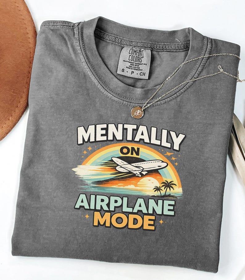 Puede incluir: Camiseta gris con la frase "MENTALLY ON AIRPLANE MODE" en blanco y amarillo. El dise&ntilde;o incluye un arco&iacute;ris, un avi&oacute;n, palmeras y una puesta de sol. Un collar plateado con un colgante est&aacute; sobre la camiseta.