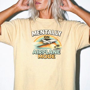 Puede incluir: Camiseta amarillo claro con la frase "MENTALLY ON AIRPLANE MODE". El dise&ntilde;o incluye un arco&iacute;ris, un avi&oacute;n, palmeras y una puesta de sol. La camiseta es de estilo casual y c&oacute;modo.