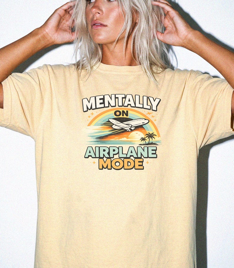 Puede incluir: Camiseta amarillo claro con la frase "MENTALLY ON AIRPLANE MODE". El dise&ntilde;o incluye un arco&iacute;ris, un avi&oacute;n, palmeras y una puesta de sol. La camiseta es de estilo casual y c&oacute;modo.