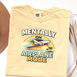 Puede incluir: Camiseta amarilla clara con el texto "MENTALLY ON AIRPLANE MODE" sobre un gr&aacute;fico de un avi&oacute;n volando sobre un arco&iacute;ris y una escena tropical. Un collar dorado con un colgante de estrella est&aacute; sobre la camiseta.