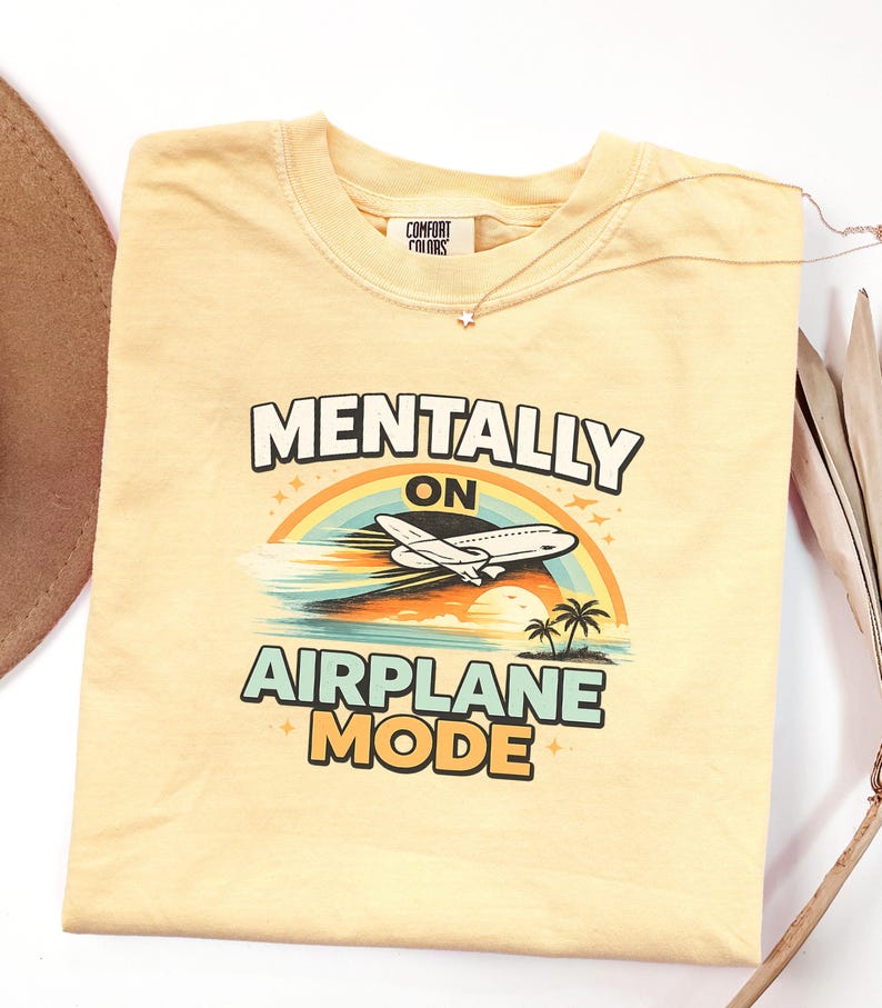 Puede incluir: Camiseta amarilla clara con el texto "MENTALLY ON AIRPLANE MODE" sobre un gr&aacute;fico de un avi&oacute;n volando sobre un arco&iacute;ris y una escena tropical. Un collar dorado con un colgante de estrella est&aacute; sobre la camiseta.