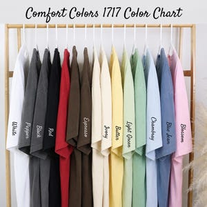 Puede incluir: Una carta de colores de camisetas Comfort Colors 1717 colgadas en un estante de madera. Las camisetas son de varios colores, incluyendo blanco, pimienta, negro, rojo, espresso, marfil, mantequilla, verde claro, chambray, azul vaquero y flor. El texto "Comfort Colors 1717 Color Chart" est&aacute; en la parte superior.