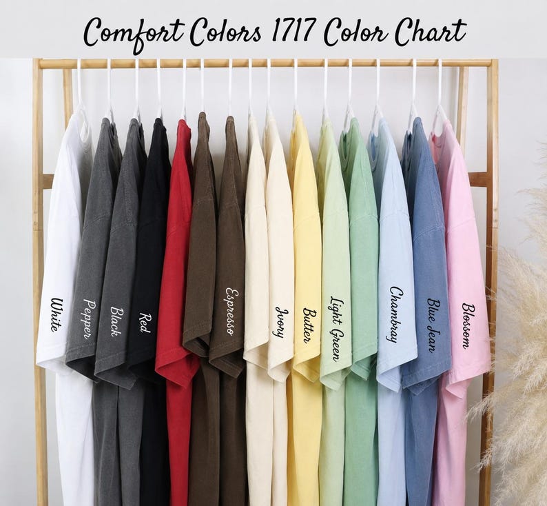 Puede incluir: Una carta de colores de camisetas Comfort Colors 1717 colgadas en un estante de madera. Las camisetas son de varios colores, incluyendo blanco, pimienta, negro, rojo, espresso, marfil, mantequilla, verde claro, chambray, azul vaquero y flor. El texto "Comfort Colors 1717 Color Chart" est&aacute; en la parte superior.
