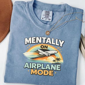 Puede incluir: Una camiseta azul con las palabras "MENTALLY ON AIRPLANE MODE" en blanco y naranja. El gr&aacute;fico presenta un arco&iacute;ris, un avi&oacute;n, palmeras y una puesta de sol. Un collar dorado con un colgante est&aacute; sobre la camiseta.