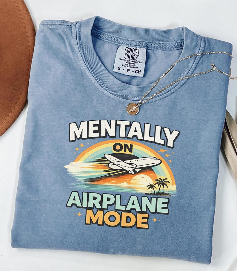 Puede incluir: Una camiseta azul con las palabras "MENTALLY ON AIRPLANE MODE" en blanco y naranja. El gr&aacute;fico presenta un arco&iacute;ris, un avi&oacute;n, palmeras y una puesta de sol. Un collar dorado con un colgante est&aacute; sobre la camiseta.