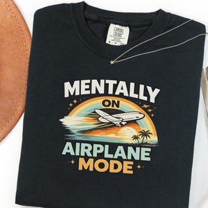 Puede incluir: Camiseta negra con las palabras "MENTALLY ON AIRPLANE MODE" en blanco y naranja. El dise&ntilde;o incluye un arco&iacute;ris, un avi&oacute;n, palmeras y una puesta de sol. Un collar dorado con un colgante de estrella est&aacute; sobre la camiseta.