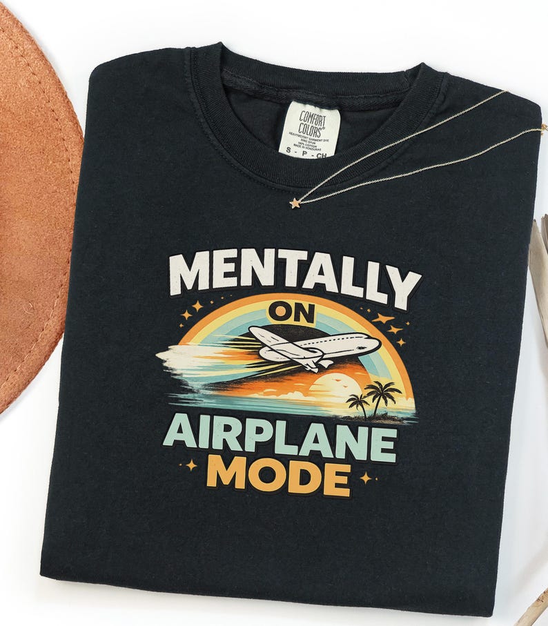 Puede incluir: Camiseta negra con las palabras "MENTALLY ON AIRPLANE MODE" en blanco y naranja. El dise&ntilde;o incluye un arco&iacute;ris, un avi&oacute;n, palmeras y una puesta de sol. Un collar dorado con un colgante de estrella est&aacute; sobre la camiseta.