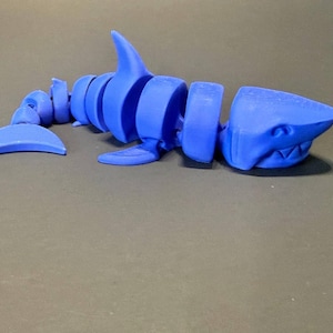 May include: A blue articulated shark toy. The toy is made of several connected segments, allowing for flexible movement. The shark features a dorsal fin, tail, and detailed head with visible teeth. The toy is designed to resemble a shark skeleton.