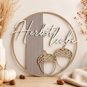 Könnte beinhalten: Ein rundes Wandbild aus Holz mit dem weißen Schriftzug "Herbstliebe", der über einem vertikalen Lamellenpaneel angebracht ist. Das Design zeigt zwei umrissene Eicheln mit Zickzackmuster auf den Kappen, eingefasst in einen dünnen Holzring.