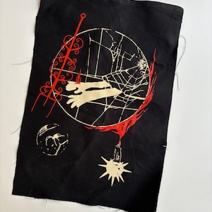 May include: Black fabric patch with a white and red graphic design. The design includes a circle with a web-like pattern, a red trident, and other stylized elements. The patch has raw edges.