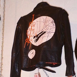May include: Black leather jacket with a distinctive back design. It showcases a large pink circle with a spiderweb pattern and a hand silhouette. There are also additional pink accents and a small circle.