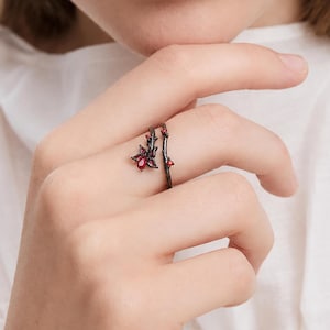 May include: An adjustable open ring featuring a dark, textured metal band designed to resemble a thin, gnarled tree branch. The ring is adorned with a small, detailed flower motif at one end and tiny red accents along the stem, set with a vibrant red stone.