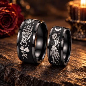 Op de afbeelding: Twee zwarte ringen met lasergegraveerde witte ontwerpen. De grotere ring toont een skeletachtige hand, een roos en kleine sterren. De kleinere ring toont een skeletachtige hand die een roos vasthoudt, een maansikkel en sterren. Beide ringen zijn glad.