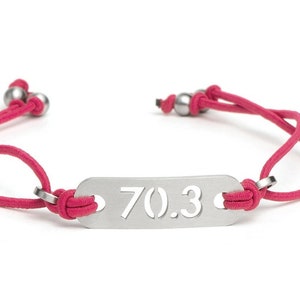 Op de afbeelding: Een roze verstelbaar koordarmband met een zilveren bedel met de tekst "70.3".