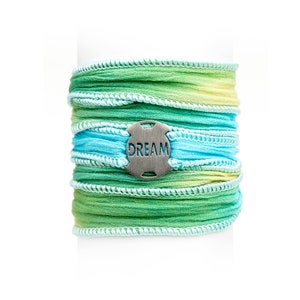 May include: A green and blue layered fabric bracelet with a silver charm that says "DREAM".