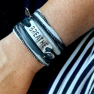 May include: A black and gray fabric bracelet with a silver charm that says "BREATHE".