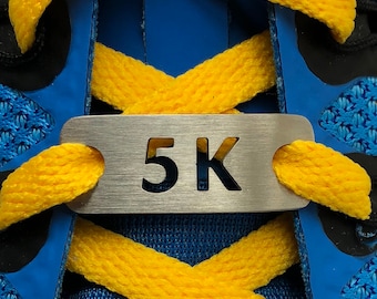 Könnte beinhalten: Ein silberner Schuhband-Anhänger aus Metall mit dem ausgeschnittenen Text "5K". Der Anhänger ist an einem Paar blauen und gelben Schnürsenkeln befestigt.