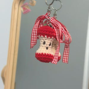 May include: Crocheted apple keychain with red and white gingham ribbon. The apple has a red top and bottom with a cream-colored center and small brown details. The keychain is attached to a silver ring.