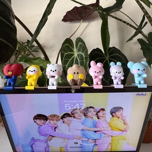 May include: A collection of seven colorful character figurines, each with unique designs and features. The figurines are arranged in a row, standing on a black surface. The background includes lush green foliage and a computer screen displaying a group of people.