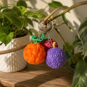 May include: Three spherical keychains featuring textured surfaces with swirling patterns. One is bright orange with a green stem, one is deep red, and one is purple with a small stem. Each keychain is attached to a silver metal ring and chain.