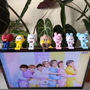 May include: Seven colourful character figurines with distinct designs and colours are displayed above a laptop screen. The screen shows a group of people. The figurines are on stands. The screen displays the date and time.