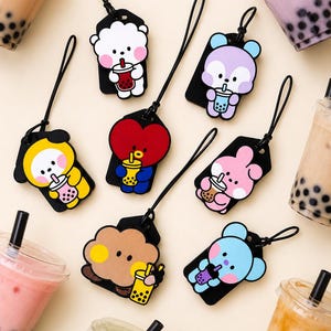 May include: A collection of seven colorful silicone charms, each featuring a cartoon character holding a bubble tea drink. The charms are attached to black cord loops and display various characters in shades of white, yellow, red, pink, brown, blue, and purple.