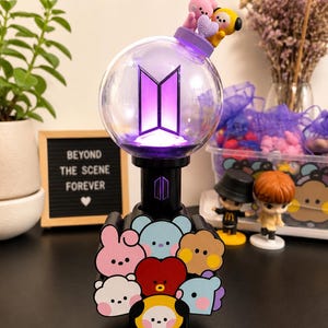 Puede incluir: Un bastón de luz BTS esférico con un interior brillante de color púrpura y un mango negro. La base está decorada con caras de personajes de dibujos animados coloridos. Pequeñas figuras de un conejo rosa y un personaje amarillo están arriba.