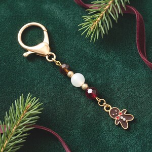 May include: A gold-toned metal keychain featuring a dangling gingerbread man charm with a red bow tie and white accents, connected to a series of beads including a large white sphere, two smaller gold spheres, and two faceted dark red beads.