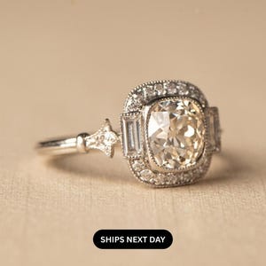 May include: A silver engagement ring with a large, square-cut, light-colored center stone. The ring features smaller stones around the center and on the band. The ring has a vintage, art deco style.