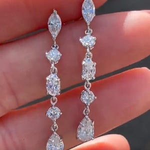 May include: Dangling earrings with multiple clear, sparkling gemstones. The earrings feature a series of various shaped stones, including marquise, round, and pear cuts, connected by delicate links. The stones are set in a silver or white metal.