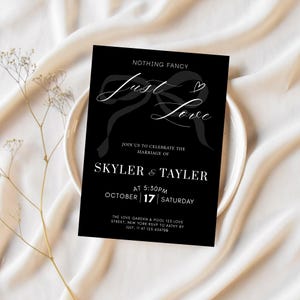 May include: A black rectangular wedding invitation card featuring white text that reads Nothing Fancy Just Love, followed by the names Skyler & Tayler, the date October 17 Saturday at 5:30pm, and a venue address with RSVP details.