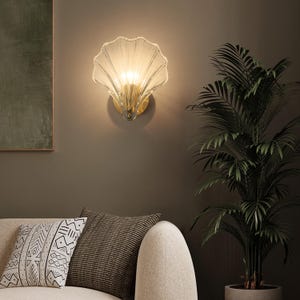 Op de afbeelding: Een decoratieve wandlamp met een schelpvormige glazen kap. De lamp heeft een goudkleurige basis en straalt een warme gloed uit. De lamp is gemonteerd op een donkere muur, met een bank en een potplant op de voorgrond.