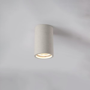 May include: A cylindrical, light-colored ceiling light fixture. The fixture has a textured surface and a recessed light source at the bottom. The light is emitting a warm, soft glow. The fixture is mounted on a white ceiling.