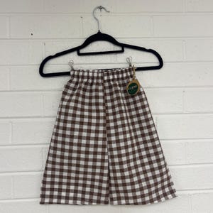 May include: Brown and white gingham shorts hanging on a black hanger against a white brick wall. The shorts have an elasticated waistband and a tag that says "CLEMI".