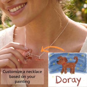 Op de afbeelding: Een zilveren ketting met twee bedels: één in de vorm van een bruine hond en een andere met de tekst "Doray" in het rood. Het ontwerp is gebaseerd op een kindertekening van een bruine hond tegen een blauwe sterrenhemel met de naam "Doray".