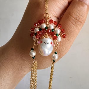 May include: A gold-colored necklace featuring a pendant with a whimsical design. The pendant includes a large, white, pear-shaped pearl, red and white beads, and gold chain tassels. The necklace is held in a hand against a neutral background.