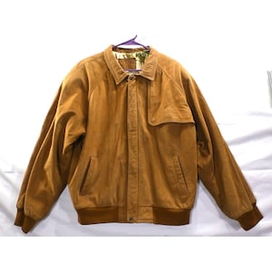 May include: A tan suede jacket featuring a collared neckline, a front button closure, and long sleeves with ribbed cuffs. The jacket includes two side pockets, a flap detail on the upper chest, and a patterned lining visible at the collar.