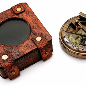 May include: A vintage-style sundial and a leather-bound box with a circular window. The sundial is made of brass with Roman numerals and a pivoting arm. The box is brown with decorative patterns and brass studs.