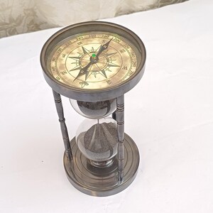 May include: A dark metallic hourglass featuring a compass integrated into the top frame. The compass dial has a cream-coloured face with black cardinal directions and degree markings. The hourglass contains dark sand within two glass bulbs.
